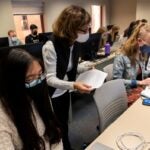 A class of students wearing face masks all sitting at desks with laptops while a female instructor walks around holding a sheet of paper and wearing glasses and a black vest over a white sweater.