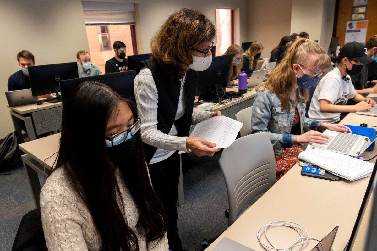 A class of students wearing face masks all sitting at desks with laptops while a female instructor walks around holding a sheet of paper and wearing glasses and a black vest over a white sweater.