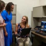 A woman with brown curly hair wearing a pink dress and a woman with short brown hair wearing a blue dress stand over a blonde woman who is seated in a chair holding a laptop and wearing a navy blue and pink floral shirt and tan pants.