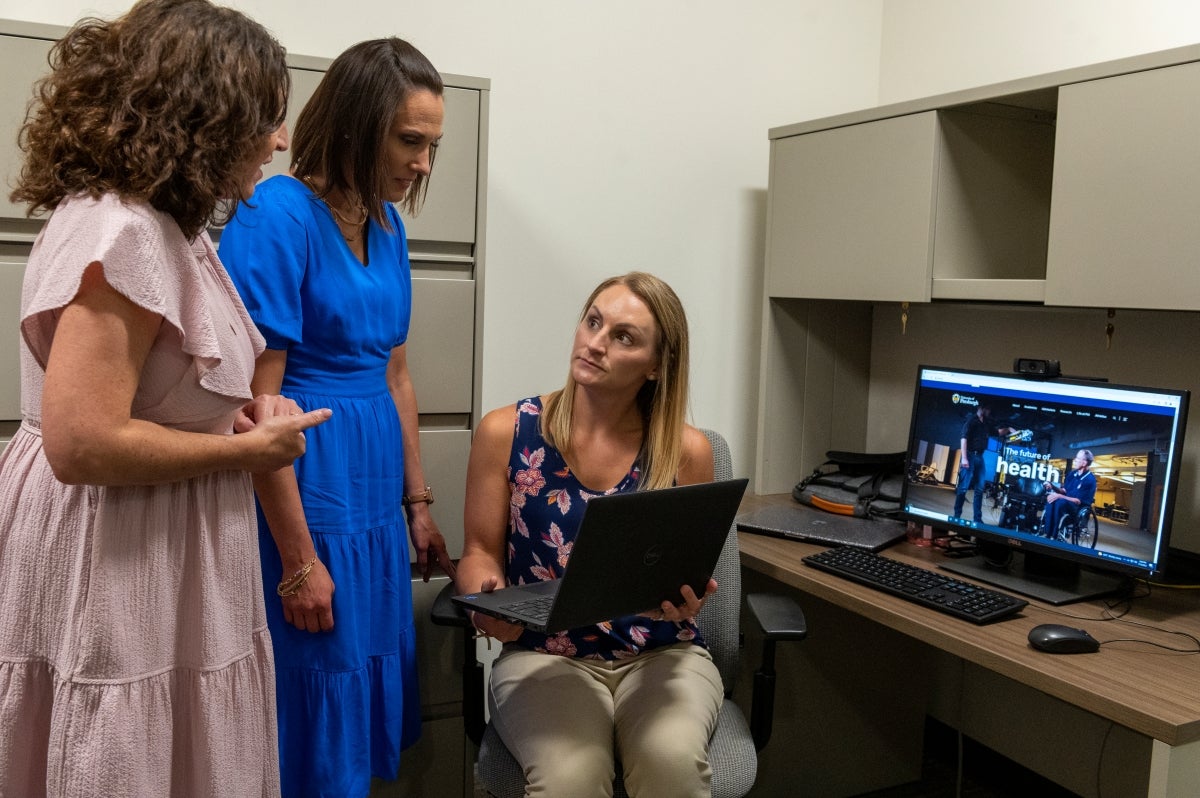 A woman with brown curly hair wearing a pink dress and a woman with short brown hair wearing a blue dress stand over a blonde woman who is seated in a chair holding a laptop and wearing a navy blue and pink floral shirt and tan pants.