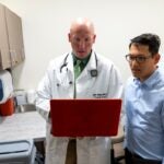 A bald man wearing a lab coat over a dark green collared shirt and a light green striped tie holds a red laptop while a man with black hair wearing a light blue collared shirt looks at the laptop.