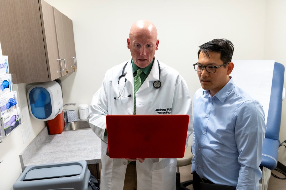 A bald man wearing a lab coat over a dark green collared shirt and a light green striped tie holds a red laptop while a man with black hair wearing a light blue collared shirt looks at the laptop.