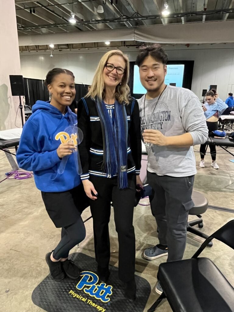 A woman with dark hair pulled back wearing a blue sweatshirt and a black skirt standing next to a woman with blonde hair wearing glasses and a blue scarf over a black and white sweater and a man with dark hair wearing a gray shirt and black pants.