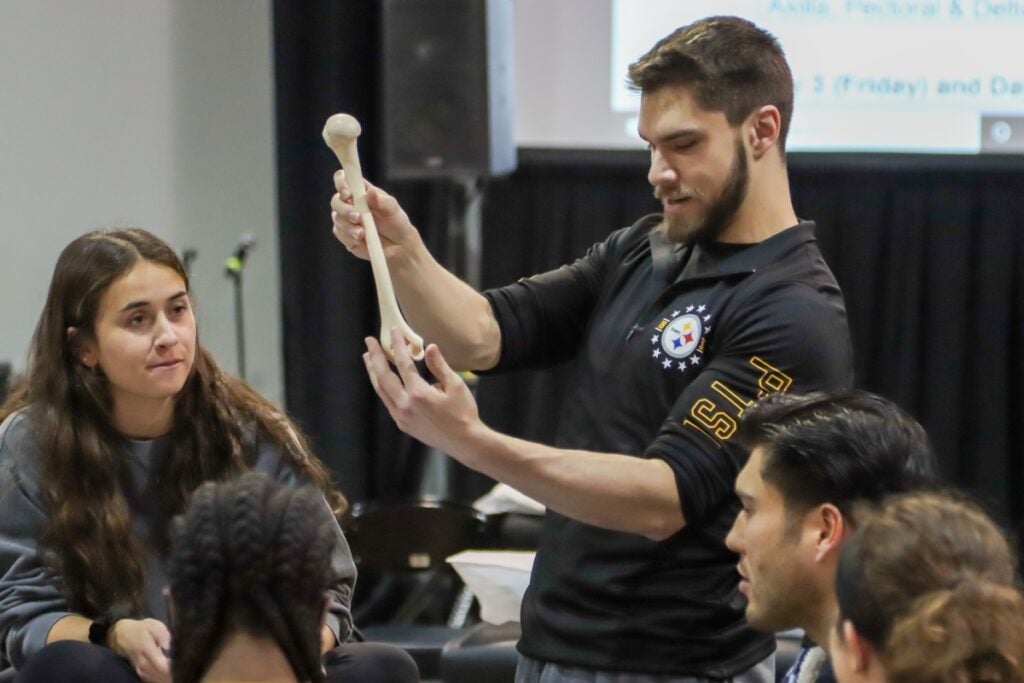 A man with brown hair wearing a black shirt holding a bone while men and women look on.