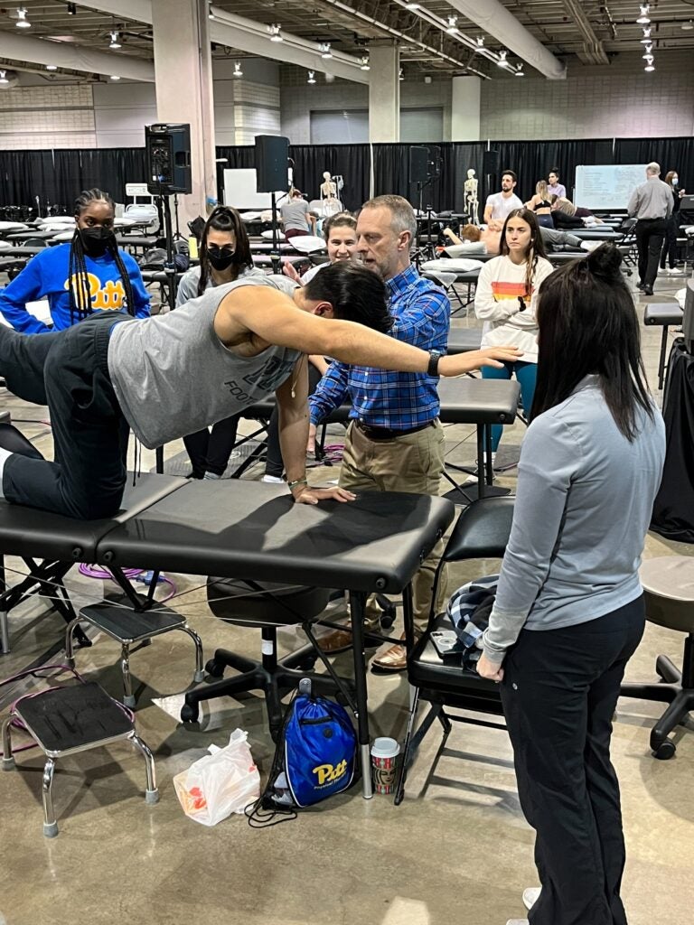 A man with gray hair wearing a blue plaid shirt and khaki pants instructing a person with dark hair wearing a gray shirt and black pants who is stretching on a table. Men and women stand in the background watching the demonstration.