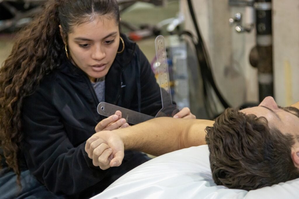 A woman with long brown hair wearing a dark sweatshirt and gold earrings holds a clear tool up against a man's arm, who has brown hair and is laying down.