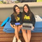 Two women with dark brown hair wearing black Pitt sweatshirts sitting together while holding laptops.