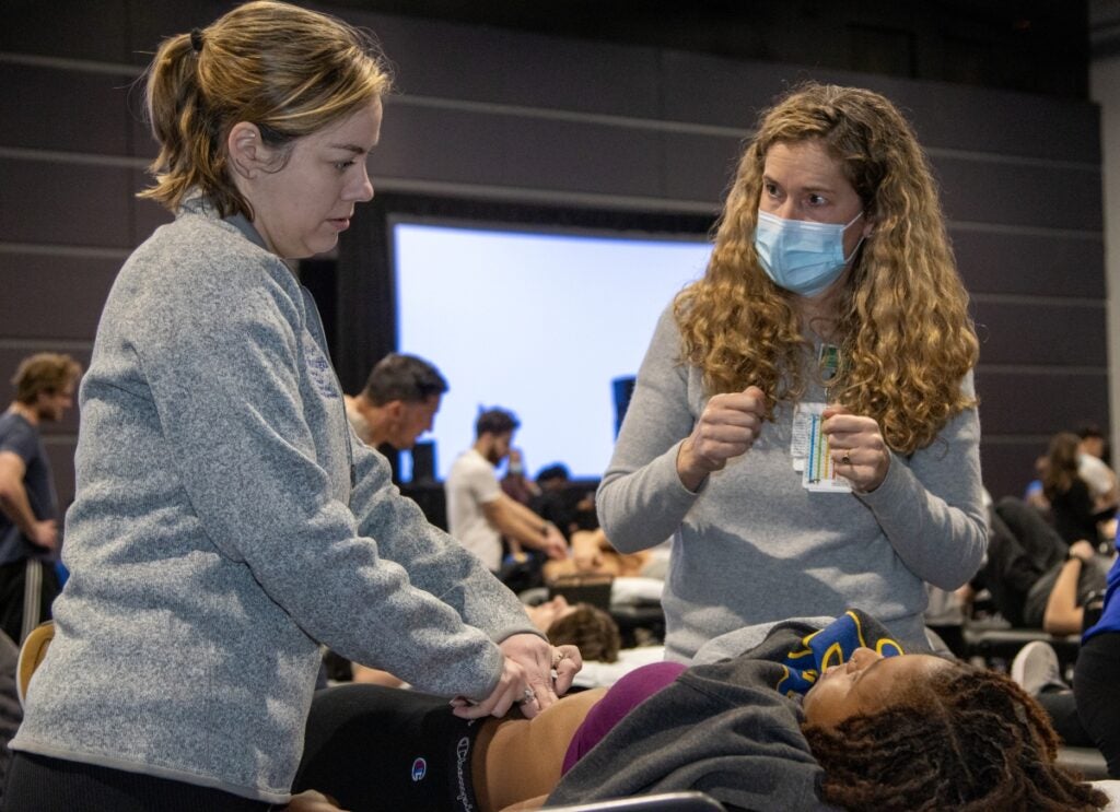 A woman with blonde curly hair wearing a gray sweater and a face mask instructs a woman with short brown hair wearing a gray sweatshirt as she exams a woman with brown braided hair.