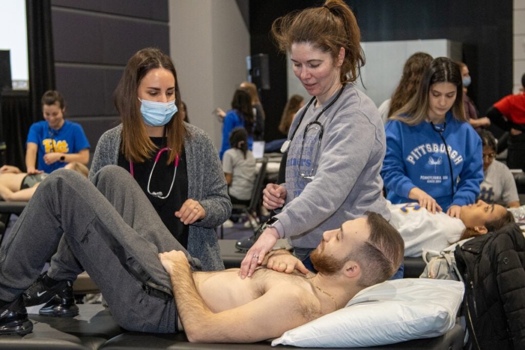 A woman with brown hair wearing a gray cardigan over a black shirt and a woman with auburn hair wearing a gray shirt stand over a man in a bed wearing gray sweatpants.