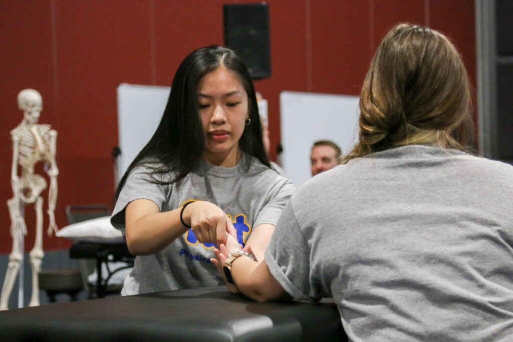 A woman with long black hair wearing a gray shirt holds the wrist of a woman with brown hair wearing a gray shirt.