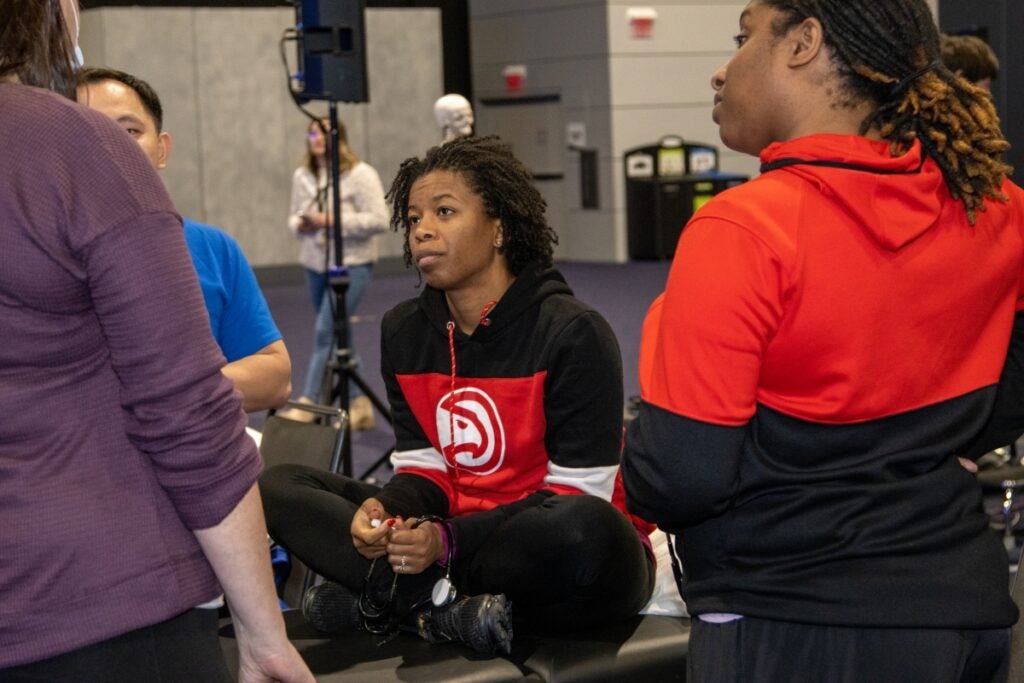 Three people stand together listening to a woman with brown hair and a purple sweater. One person, wearing a black and red sweatshirt sits on a table.