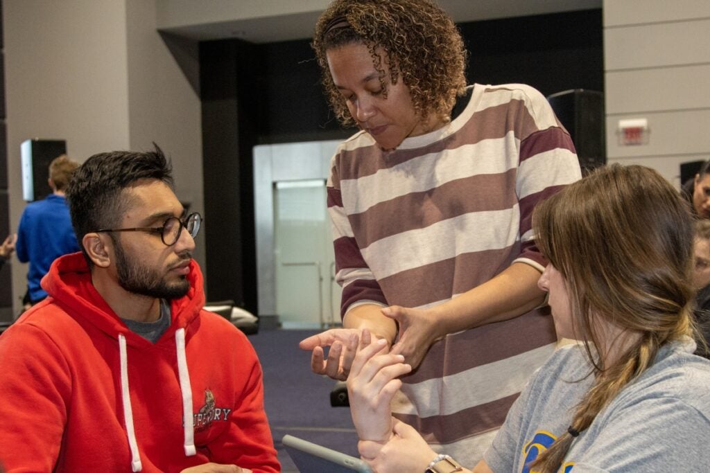 A woman with short brown curly hair wearing a brown and white striped shirt instructs a man with black hair wearing a red sweatshirt and a woman with brown hair wearing a gray shirt.