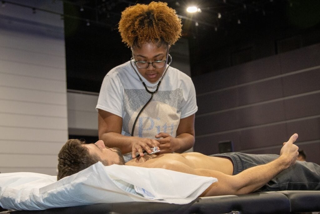 A woman with auburn braided hair wearing a white shirt using a stethoscope on a man with brown hair wearing gray sweatpants.