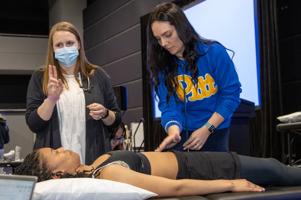 A woman with light brown hair wearing a gray cardigan over a white dress instructs a woman with black hair wearing a blue sweatshirt while she uses a stethoscope to examine a woman with black braids lying on a table.