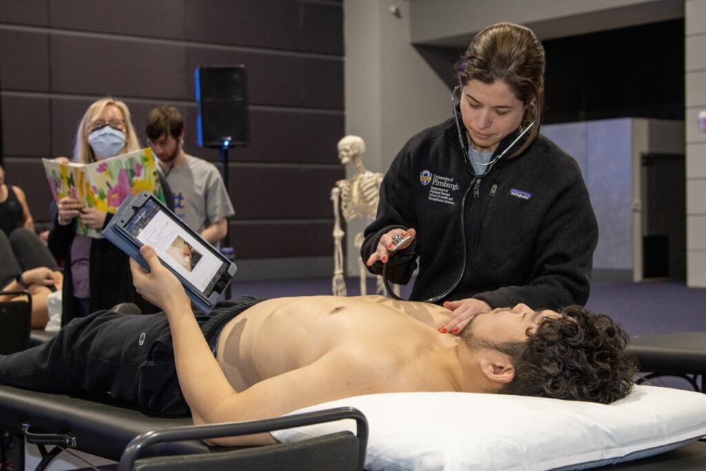 A woman with brown hair wearing a black zip-up uses a stethoscope on a male with brown curly hair who is lying on a bed wearing black pants and holding a tablet.