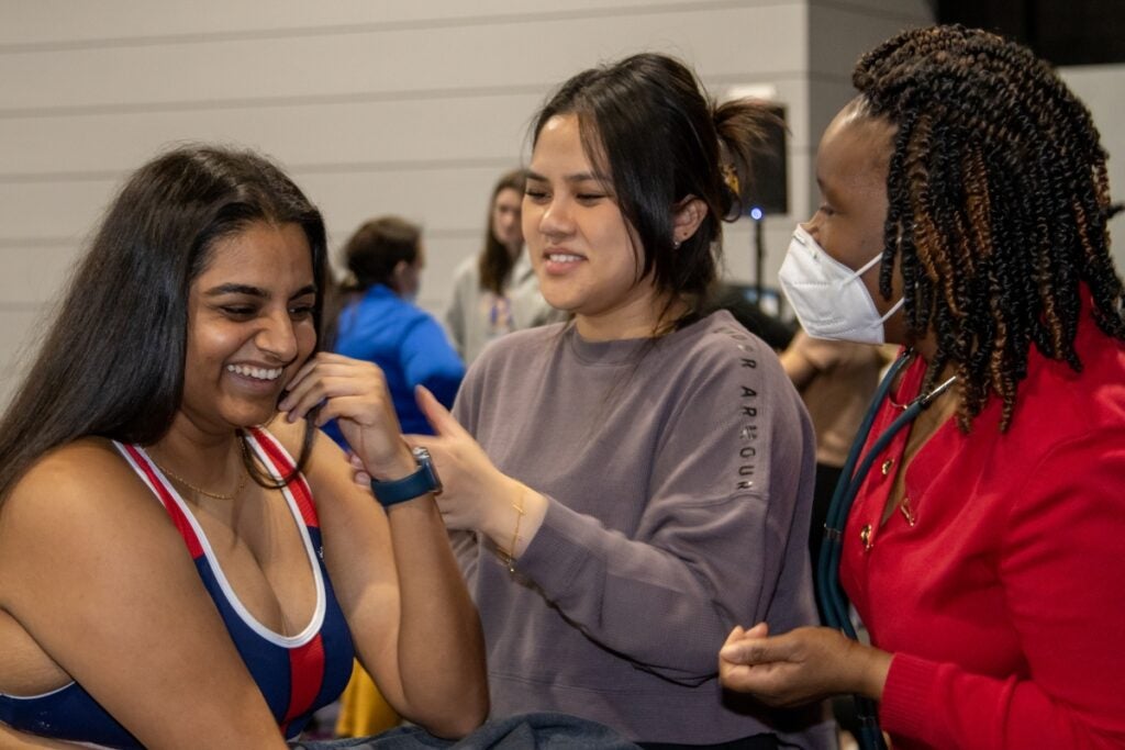 A woman with long brown hair wearing a navy and red sports bra, a woman with black hair pulled back wearing a brown sweatshirt and a woman with dark brown braids wearing a red sweater, a face mask and a stethoscope all stand together talking.