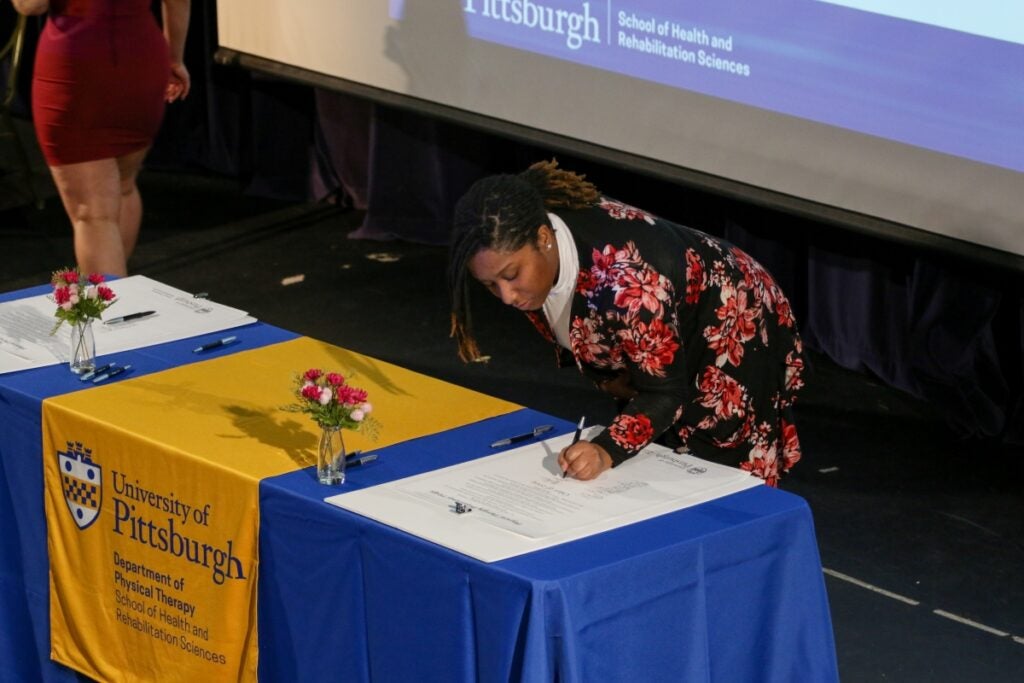 A woman with black and brown braided hair wearing a pink floral and black dress over a white turtleneck standing over a table with blue tablecloths signing a paper.