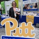 A woman with short curly hair wearing a black cardigan over a white blouse with peach and black detailing who stands behind a Pitt OT sign.