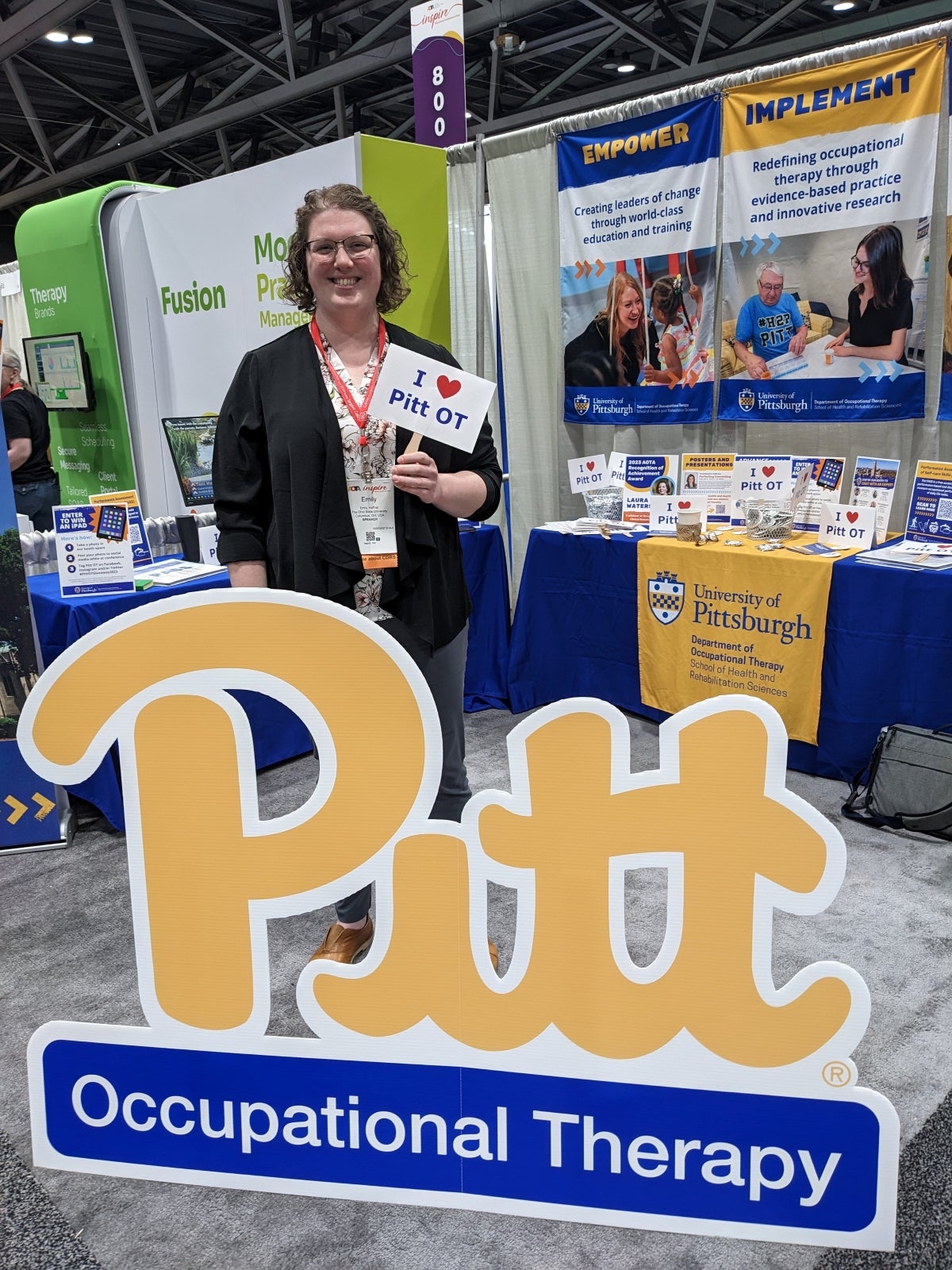 A woman with short curly hair wearing a black cardigan over a white blouse with peach and black detailing who stands behind a Pitt OT sign.