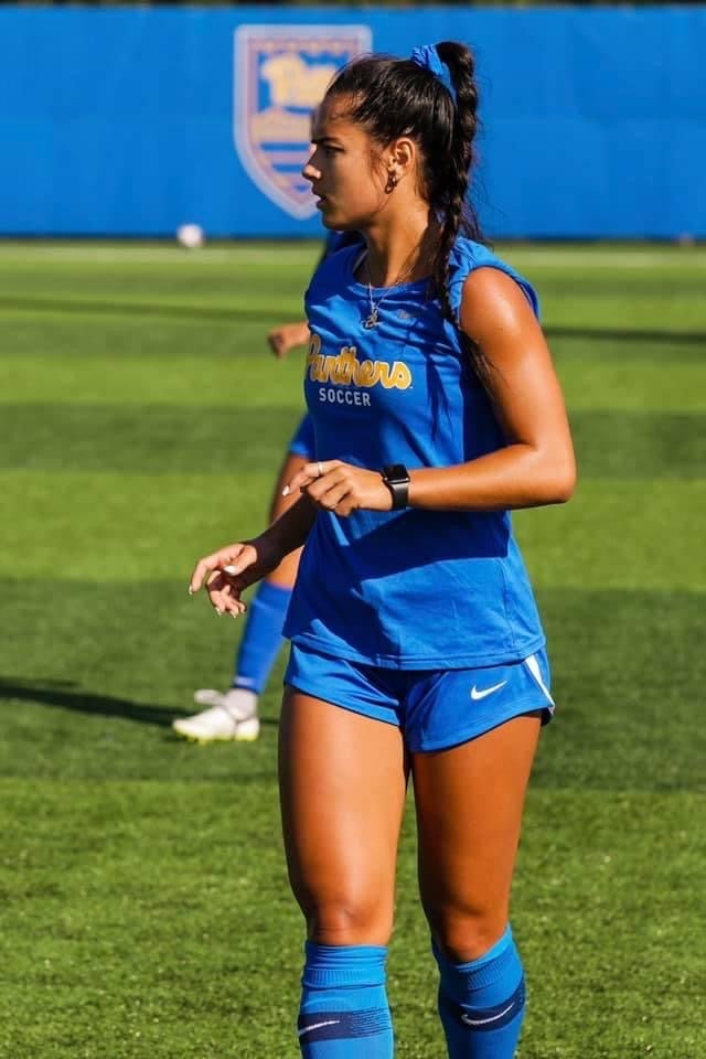 A woman with dark brown hair in a braid wearing a blue Pitt soccer tank top, blue shorts and blue socks while on a soccer field.