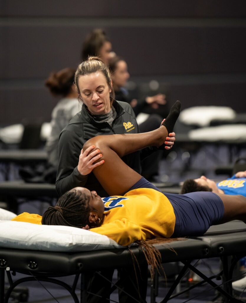 A woman with short brown hair pulled back wearing a black zip-up performs a physical therapy exercise on a woman with long dark brown braids wearing a yellow sweatshirt while lying on a table.