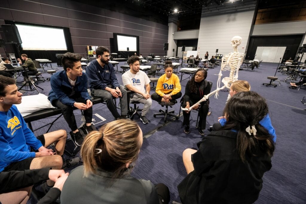 A group of men and women sitting in a circle listening to a woman with dark brown braids who is speaking while holding a skeleton model.