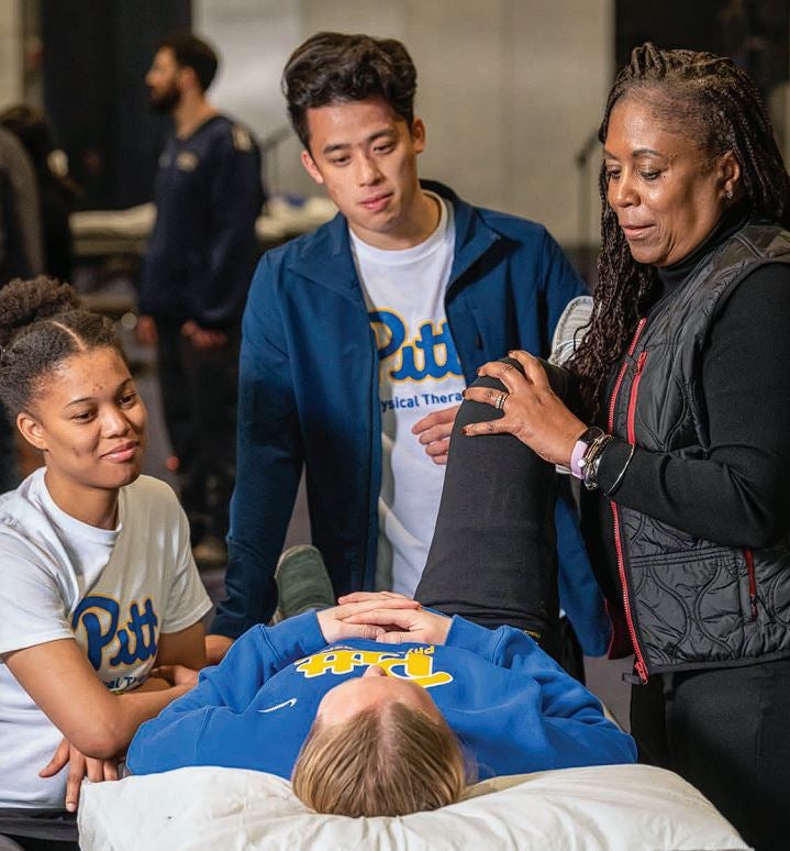 A woman with long brown braided hair wearing a black vest instructs a male and female student while examining a female student who is lying down.