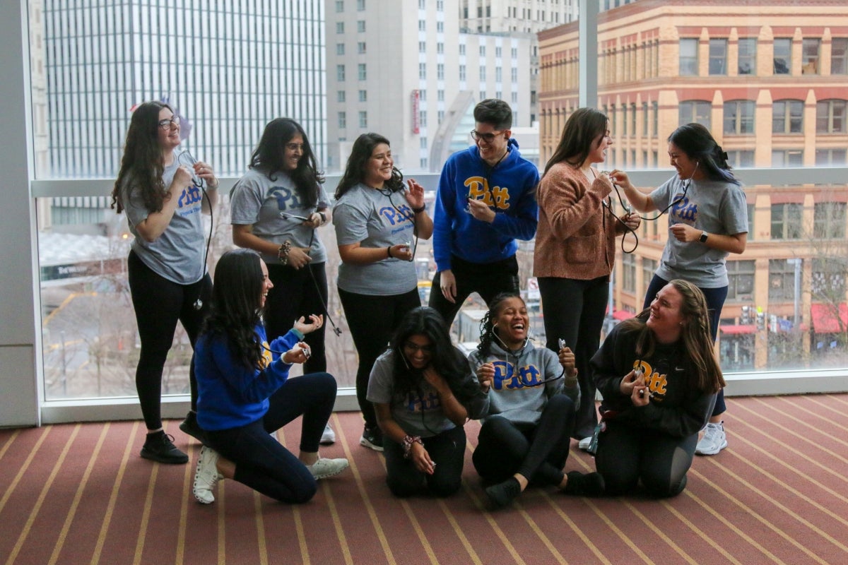 A group of men and women standing together laughing while wearing Pitt gear and holding stethoscopes.