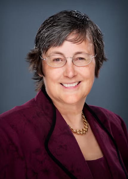 Woman with short black hair wearing silver rimmed glasses and a marron suit jacket over a maroon top with a gold necklace.