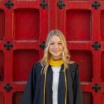 Woman with blonde hair wearing a black graduation gown and a gold graduation cord over a white dress.