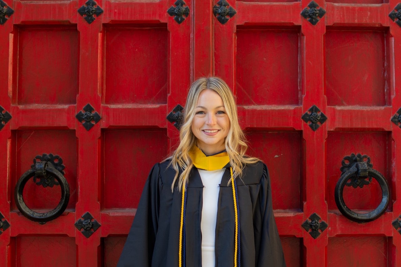 Woman with blonde hair wearing a black graduation gown and a gold graduation cord over a white dress.