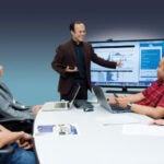 A man wearing a brown suit stands at the front of a room pointing at a screen with data on it, while four people sit around a table looking at him.