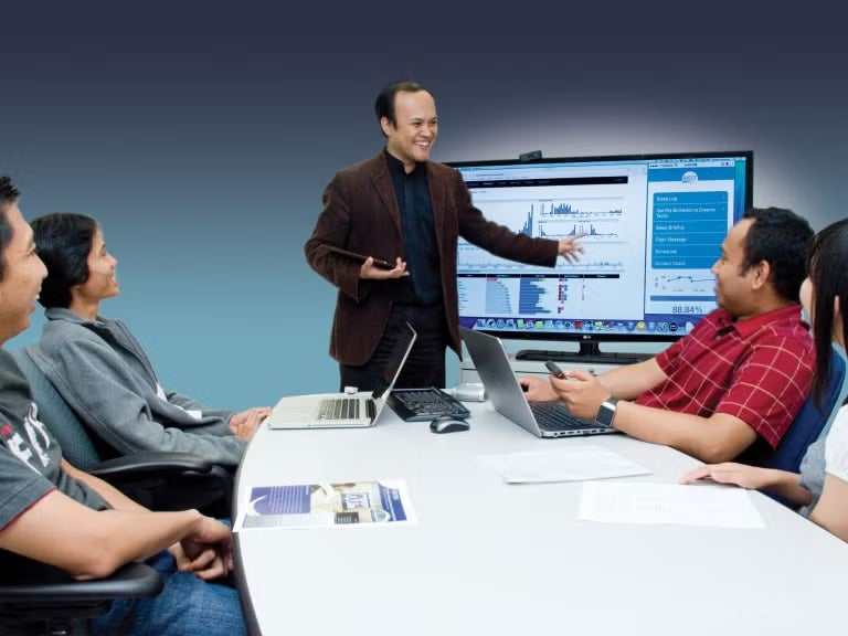 A man wearing a brown suit stands at the front of a room pointing at a screen with data on it, while four people sit around a table looking at him.