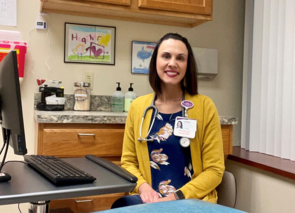 A woman with short, dark brown hair wearing a yellow cardigan over a navy blue blouse with pink and gold flowers sitting on a chair next to a countertop and a desktop computer.