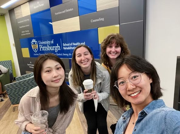 Four women standing together smiling, two of which are holding coffees.