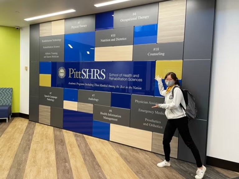 Woman wearing a white sweatshirt, black jeans, white shoes and a backpack stands next to a colorful wall with Pitt SHRS written on it.