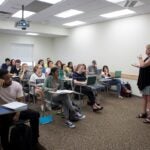 A classroom of students sitting at desks with laptops and notebooks all looking toward a woman standing at the front of the classroom who is blonde and is wearing a black cardigan over a black and white dress.