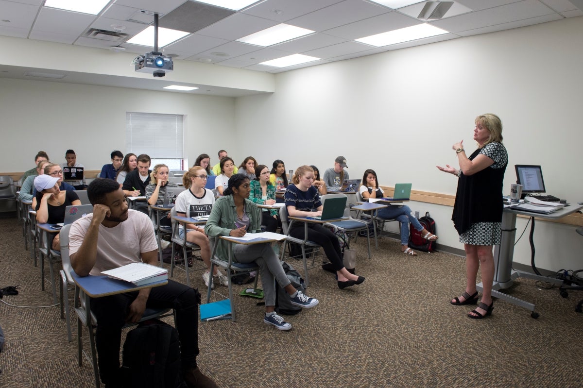 A classroom of students sitting at desks with laptops and notebooks all looking toward a woman standing at the front of the classroom who is blonde and is wearing a black cardigan over a black and white dress.