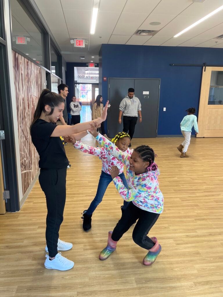 A woman and two young girls with their arms outstretched standing together smiling in a room with wood floors. The woman is wearing all black and the young girls are wearing tie-dyed sweatshirts.