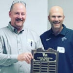 A bald man with glasses wearing a light green collared shirt holding a plaque next to a bald man wearing glasses and a dark green collared shirt.