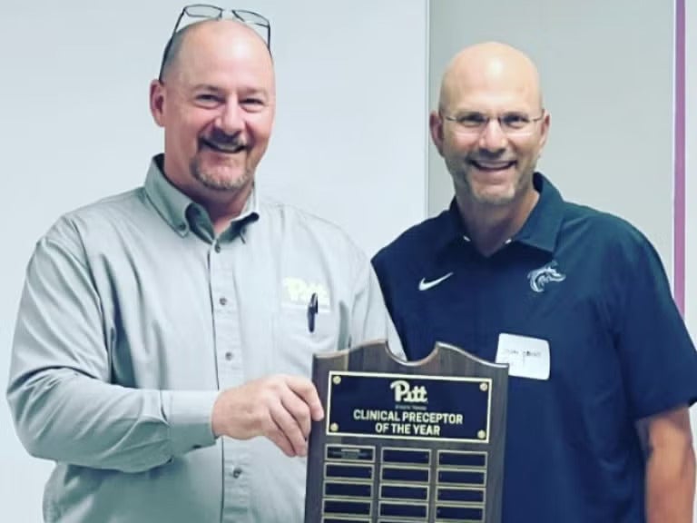 A bald man with glasses wearing a light green collared shirt holding a plaque next to a bald man wearing glasses and a dark green collared shirt.