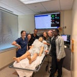 Men and women standing together around a stretcher where a training manikin lays.