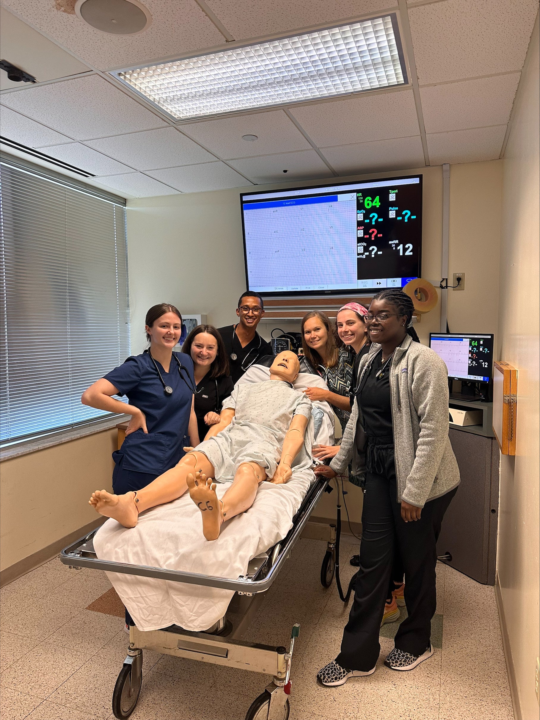 Men and women standing together around a stretcher where a training manikin lays.