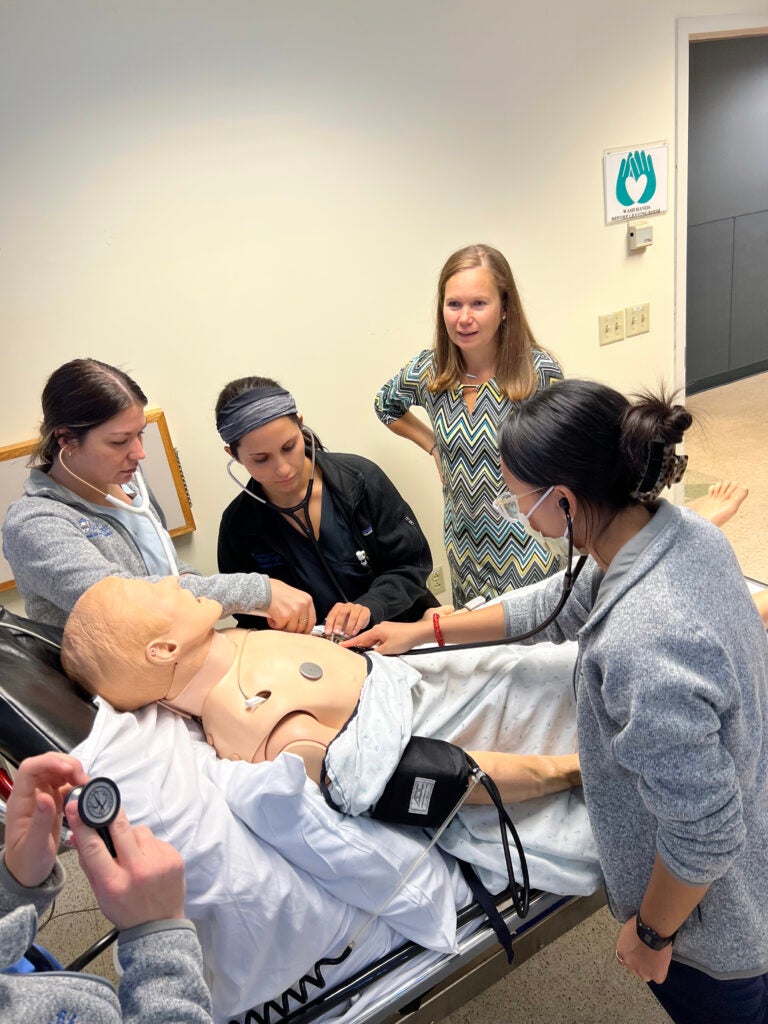 A woman with dark blonde hair waring a striped dress instructing three women wearing scrubs and stethoscopes as they stand over a training manikin.