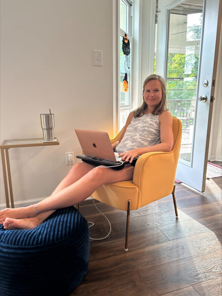 A woman with dark blonde hair wearing a gray and white camo tank top while sitting on a yellow chair with a pink laptop on her lap.