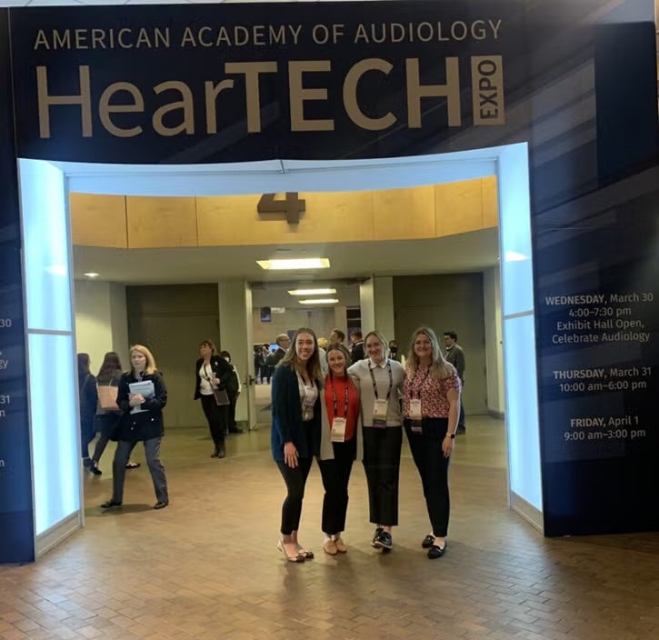 Four women standing together wearing lanyards in a hallway with a sign above them that reads "American Academy of Audiology HearTech Expo."