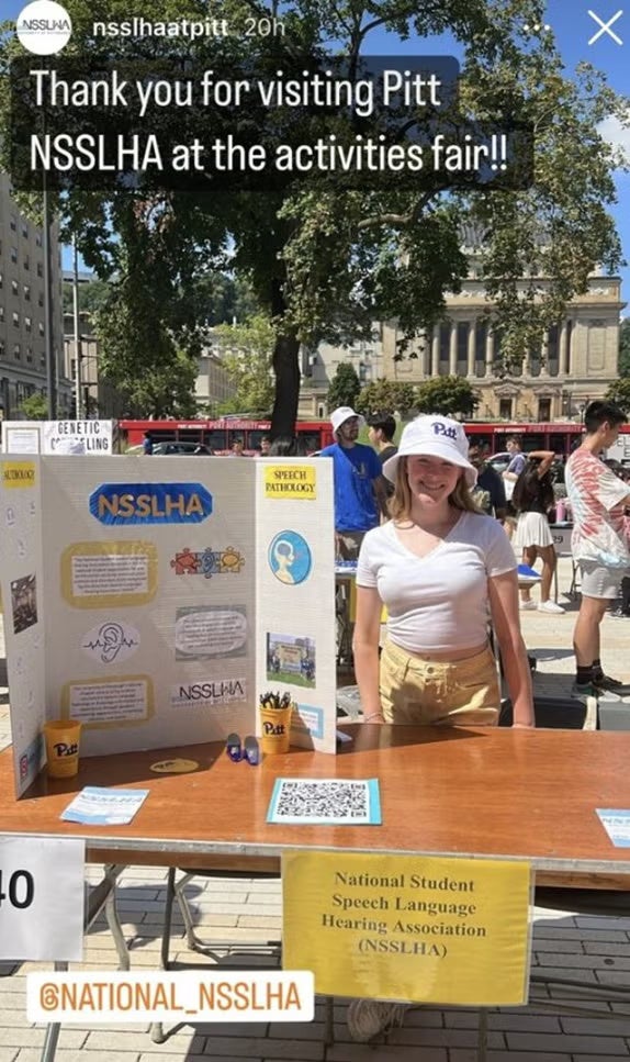 A woman with blonde hair wearing a white shirt, yellow pants and a white hat standing at a table with a poster.