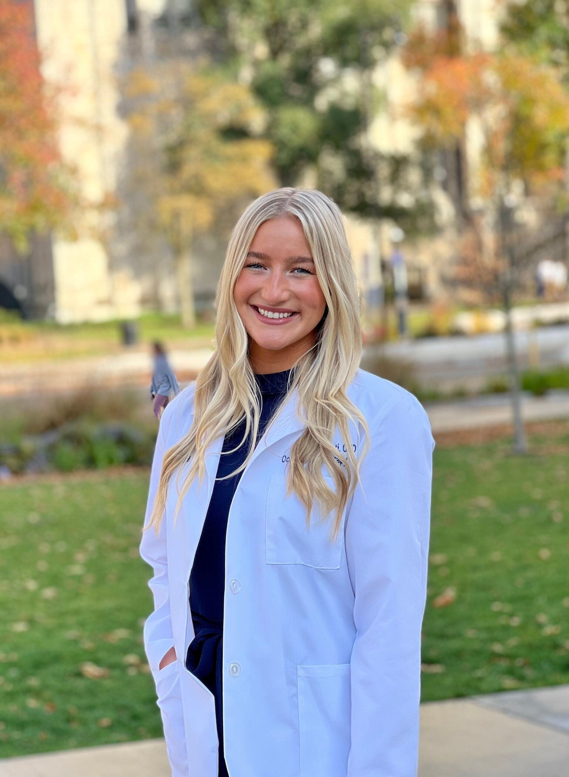 A woman with long blonde hair wearing a white lab coat over a black top.