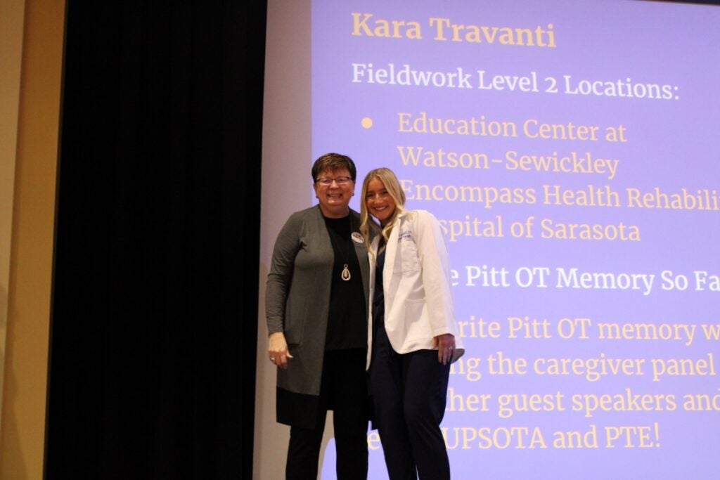 A woman with short brown hair wearing a gray cardigan over a black sweater and black pants and a woman with blonde hair wearing a white lab coat over a black top and black pants standing together in front of a projected screen.