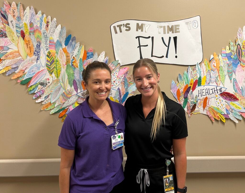 A woman with brown hair pulled back wearing purple scrubs and a woman with blonde hair pulled back wearing black scrubs standing next to each other.